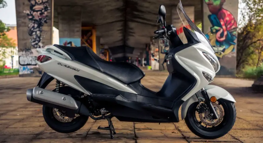 Biały skuter Suzuki Burgman 125, z czarnym siedzeniem, zaparkowany pod mostem z kolorowymi graffiti. Idealny do miasta, sprawdź suzuki burgman 125 dane techniczne.