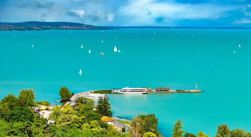 Turkusowe wody jeziora Balaton, pełne żaglówek i statków, z zielonymi wzgórzami w tle.
