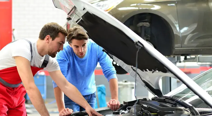 Zwei Männer blicken in den offenen Motorraum eines Autos. Der Mechaniker in Rot erklärt dem Kunden, welche Werkstatt mitgebrachte Teile einbaut.