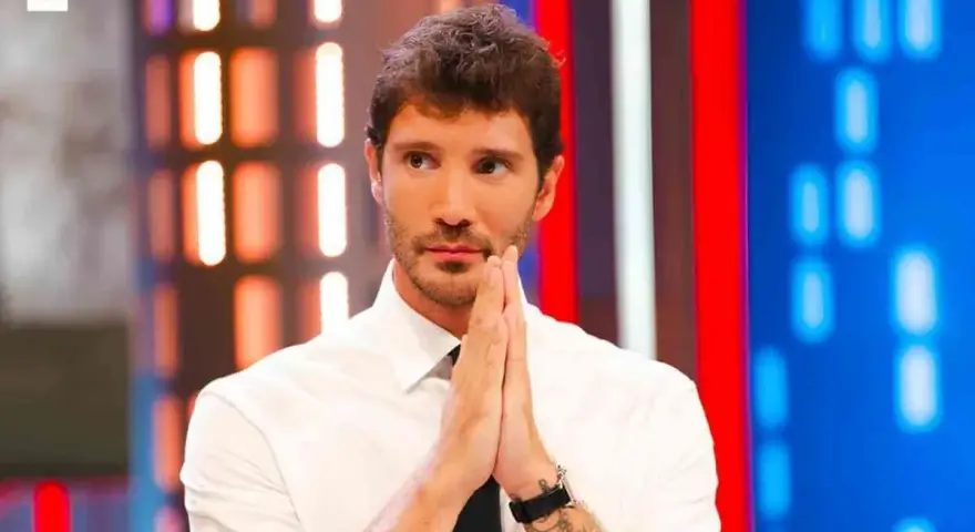 Santiago de Martino, con le mani giunte in preghiera, guarda intensamente verso l'obiettivo, con uno sfondo di luci colorate.