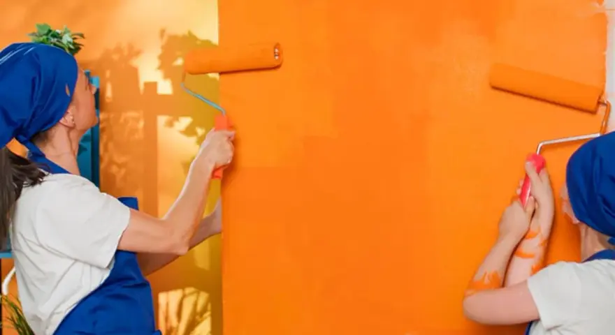 Dos personas pintan una pared de naranja brillante con rodillos. La pintura mural exterior cobra vida con vibrantes tonos anaranjados.