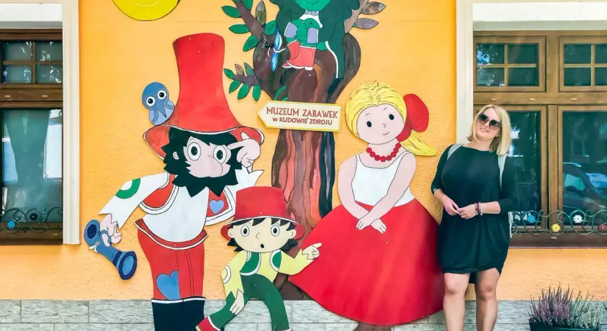Kobieta pozuje przy kolorowej ścianie Muzeum Zabawek w Kudowie-Zdroju, Dolny Śląsk. Wesołe postacie z bajek towarzyszą jej w radosnej scenerii.