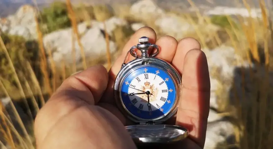 Mano sostiene un reloj de bolsillo antiguo con esfera azul y números romanos, útil para orientarse con reloj de agujas en un paisaje montañoso.