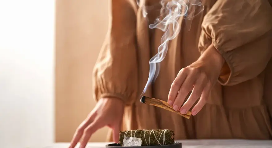 Dym z kadzidła unosi się, tworząc mistyczną atmosferę. Ręce trzymają drewno, symbolizując rytuały związane z magią i okultyzmem.
