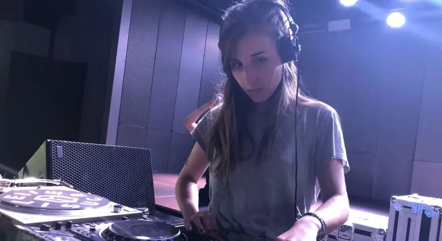 Une dj fille concentrée aux platines, casque sur les oreilles, dans une ambiance tamisée avec des lumières bleues.