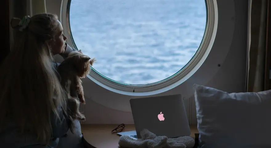 Dziewczyna z psem patrzy przez okrągłe okno na morze. Obok leży laptop z podświetlonym logo, jak prom nova star.
