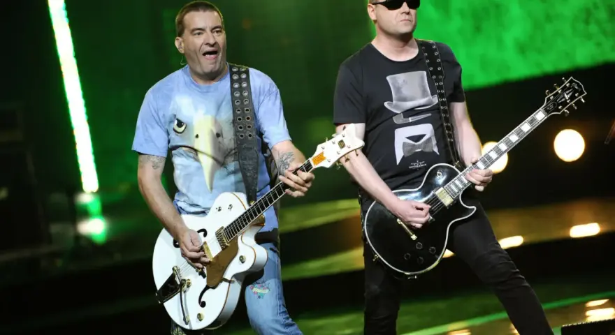 Dwóch członków Kobranocki gra na gitarach podczas koncertu. Jeden w koszulce z orłem, drugi w czarnej z cylindrem i muszką.