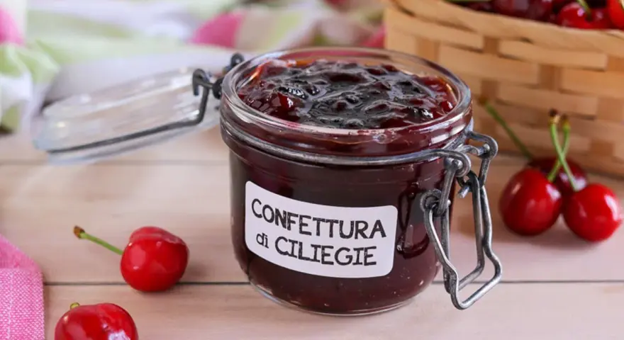Un vasetto di marmellata di ciliegie fatta in casa, con ciliegie fresche e un cestino sullo sfondo.