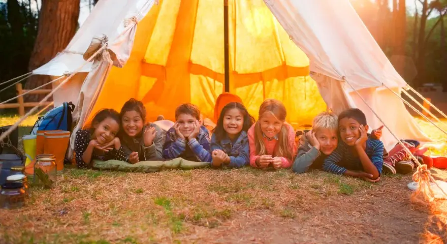 Sept enfants souriants allongés devant une tente de camping éclairée par le soleil.