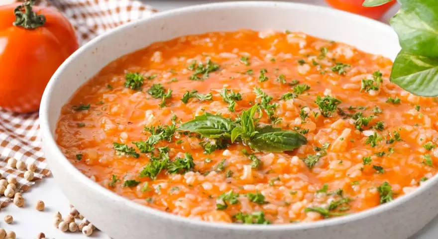 Eine cremige Tomatensuppe mit Reis, garniert mit frischer Petersilie und Basilikum. Daneben liegen frische Tomaten und Gewürzkörner.