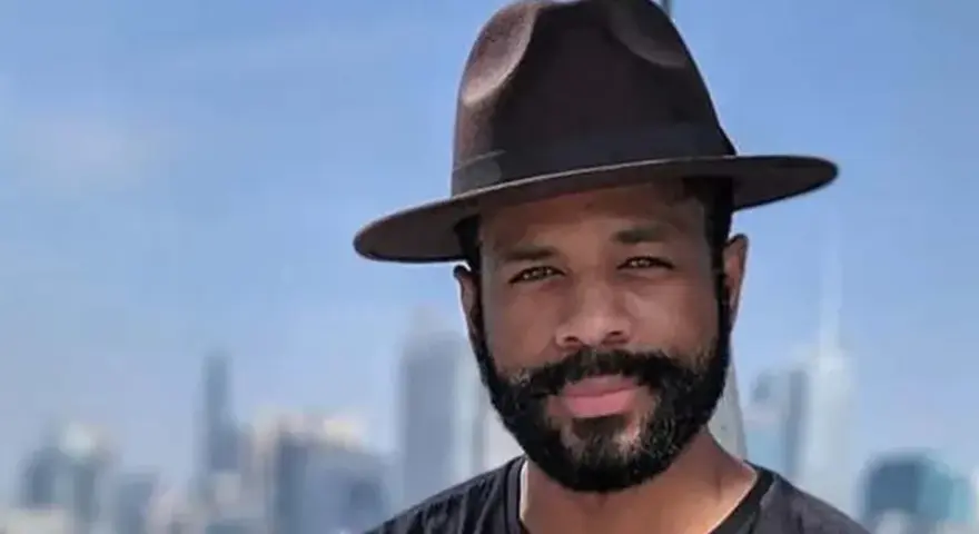 Hombre con sombrero y barba espesa, sonriendo ante un fondo de edificios y cielo azul.