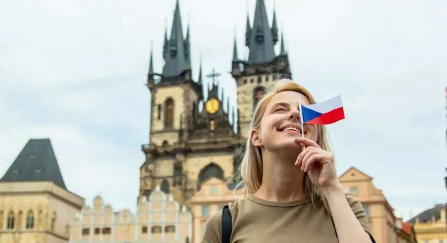 Uśmiechnięta blondynka z flagą Czech zasłaniającą oko, marzy o wysokich zarobkach w Czechach. W tle praskie kamienice i kościół.