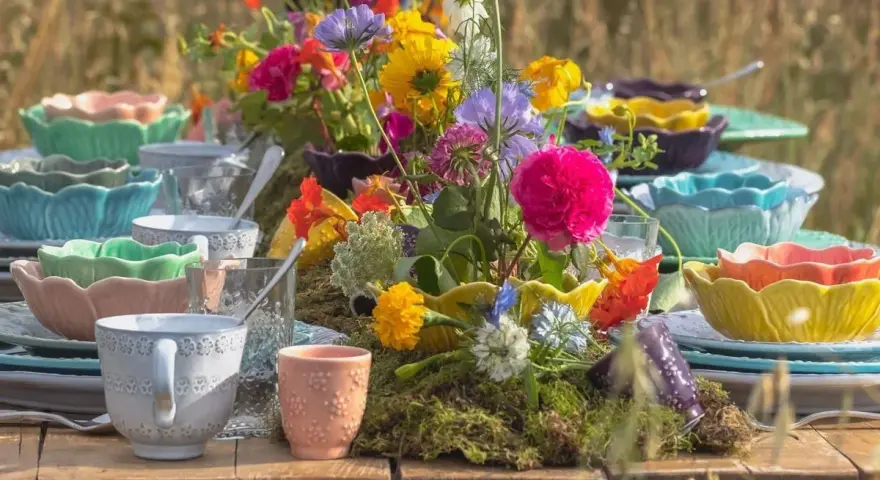 Färgglada uppläggningsfat i plast, staplade med blommor och glas, skapar en festlig dukning utomhus.