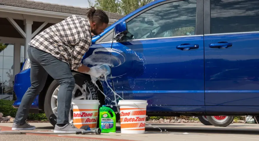 A man washes a blue car in the sun, with buckets and car wash soap nearby. It's a good day to wax a car in the sun.