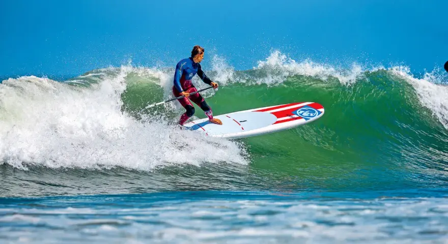 Ein Mann im Neoprenanzug surft auf einem SUP-Board eine grüne Welle. Das SUP-Wellenreiten erfordert Balance und Geschick.