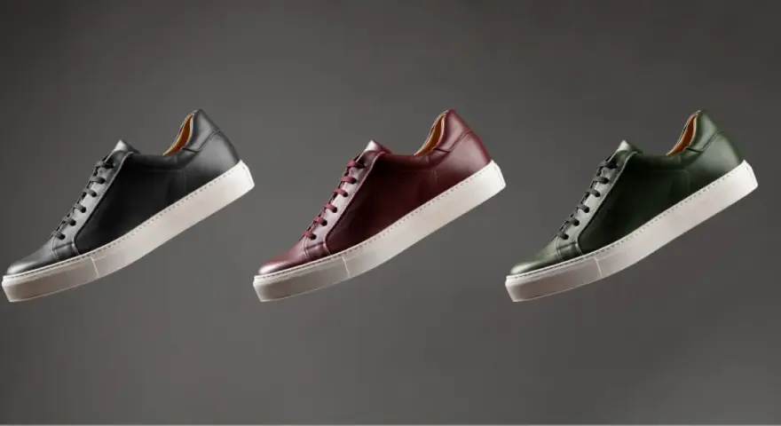 Sneakers marche famose in pelle nera, bordeaux e verde, con suola bianca, fluttuano su sfondo grigio.