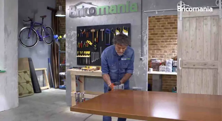 Hombre lijando mesa de madera. El taller tiene herramientas colgadas y una bicicleta morada. La madera hinchada por humedad se repara.