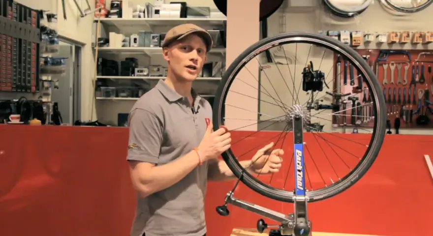 Man i keps visar hur man rikta cykelhjul i en verkstad. Verktyg hänger på väggen bakom.