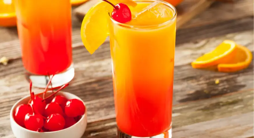Ein erfrischender Tequila Sunrise mit Orangenscheibe und Kirsche, daneben eine Schale mit Kirschen.