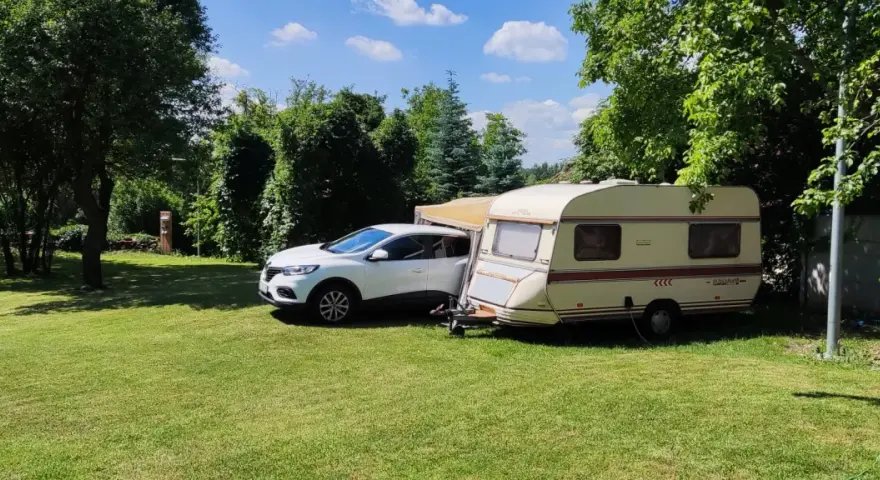 Biały samochód i przyczepa kempingowa na zielonej trawie, otoczone drzewami. Idealne miejsce na **camping smok**.