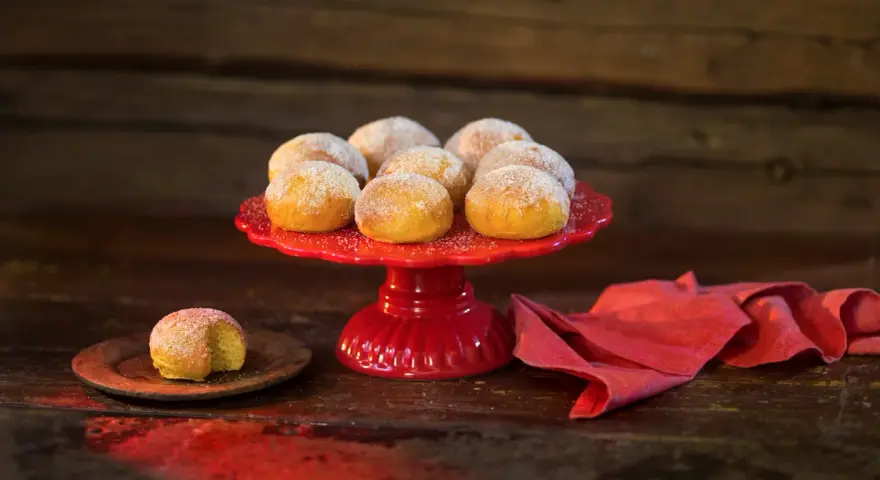 En röd tårtfat med lussebullar med kvarg, pudrade med socker. En ensam bulle ligger på en träplatta.