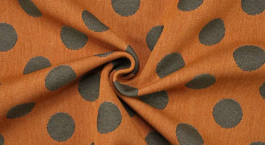 Orangefarbener Stoff mit dunklen Punkten, die wie kleine Kreise aussehen. Das Muster ist unregelmäßig und wirkt lebendig.