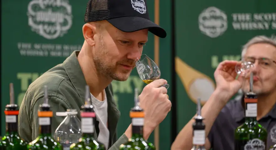 Mann mit Kappe riecht an einem Whiskyglas bei einer Whisky Messe. Flaschen stehen auf dem Tisch.