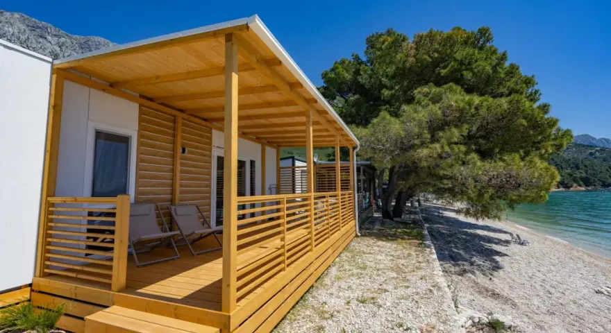 Domek mobilny z drewnianym tarasem i widokiem na morze w camp Dole Živogošće. Dwa leżaki czekają na plaży.