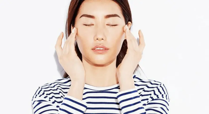A woman with closed eyes performs a gentle face massage for puffiness, her fingers delicately touching her temples.