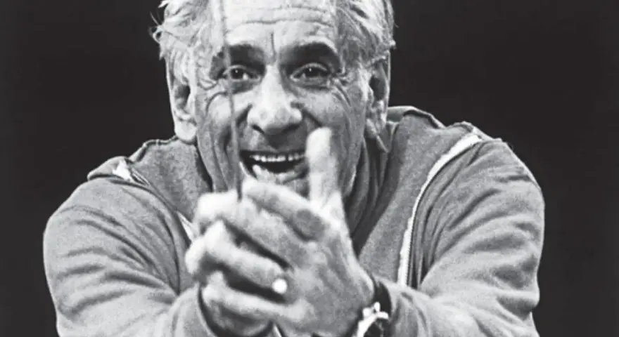 Uśmiechnięty Leonard Bernstein, dyrygent, z dłońmi złożonymi przed sobą, wpatruje się w widza.