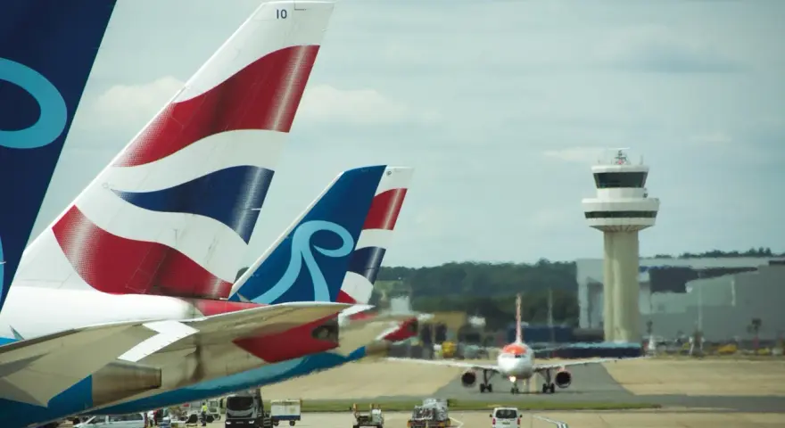 Samoloty z charakterystycznymi ogonami British Airways i innym logo czekają na start na lotnisku Londyn Gatwick. W tle wieża kontroli lotów.