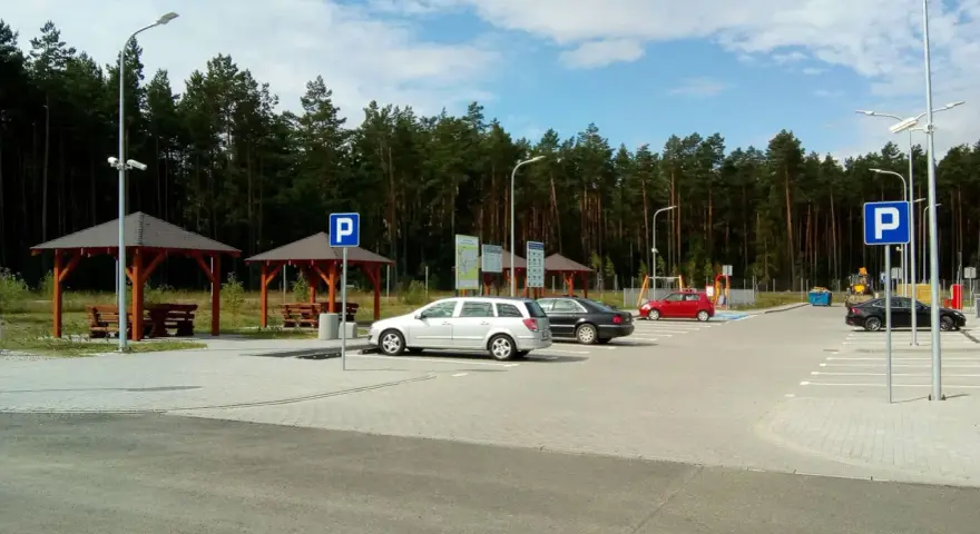 Parking przy lesie, z wiatami i ławkami. W tle las, a na parkingu kilka samochodów. To idealne miejsce na postój w drodze, może nawet w pobliżu **Orlen Sople**.