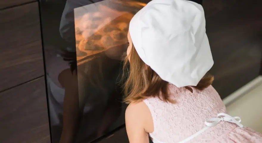 Niña con gorro de chef mirando dentro de un horno encendido.