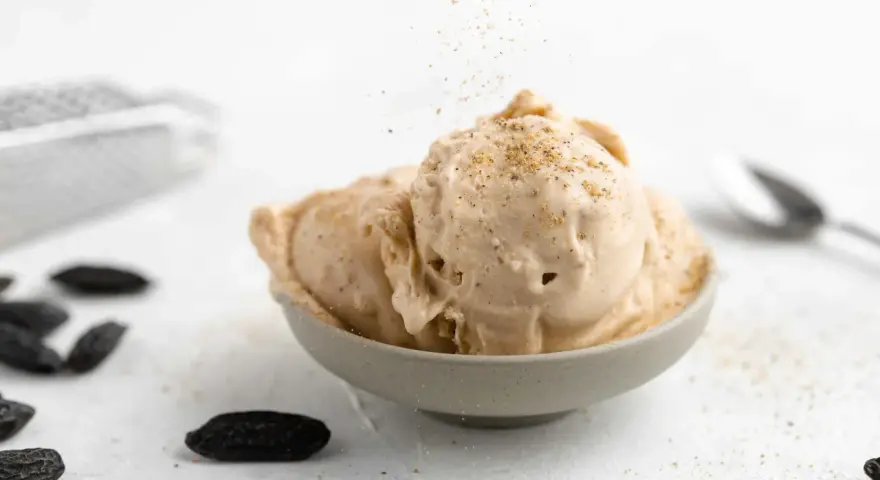 Cremiges Tonkabohnen Eis in einer Schale, bestreut mit gemahlenen Tonkabohnen. Mehrere ganze Tonkabohnen liegen daneben.