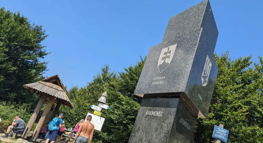 Trójstyk granic Bieszczady: monumentalny obelisk z herbami Słowacji i Ukrainy, obok drewniana wiata i turyści.