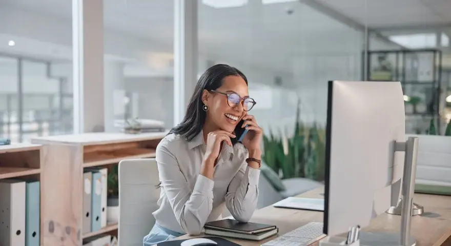 Donna sorridente al telefono in ufficio, mentre lavora al computer. Potrebbe chiedersi quanto guadagna una segretaria al mese.