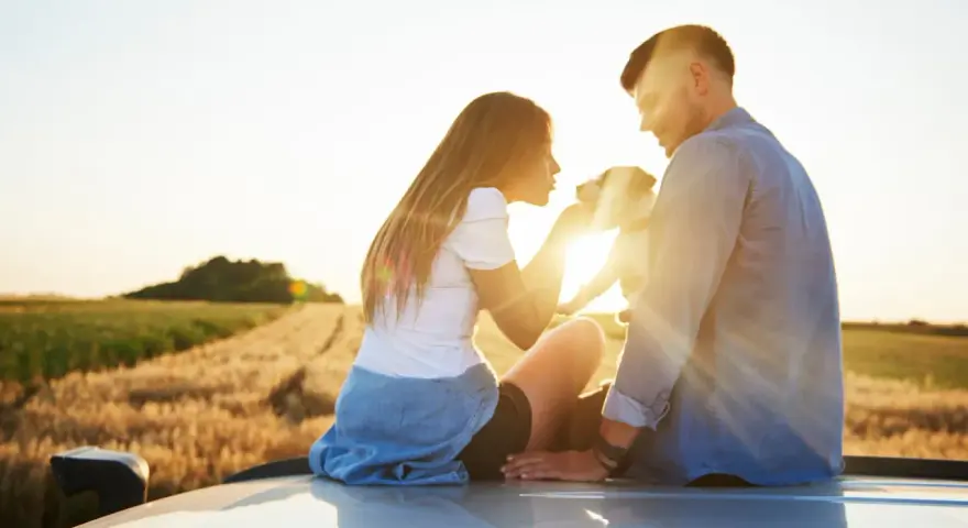 Ein Paar sitzt auf einem Auto im Sonnenuntergang, ein Hund zwischen ihnen. Sie scheinen in einer stillen Partner Meditation zu sein, umgeben von Feldern.