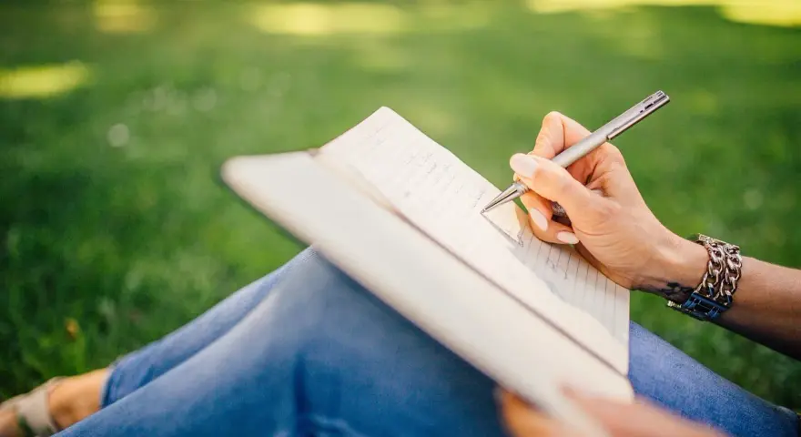 Dłoń pisze w notesie, tworząc tekst. W tle zielona trawa, a na nadgarstku bransoleta. Dopełniacz w zdaniu, jak myśli w głowie.