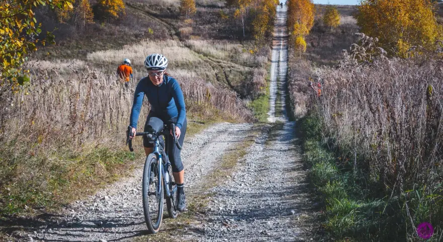 Kobieta na rowerze pokonuje kamienistą drogę w Dolinkach Krakowskich. Jesienne liście i wysoka trawa tworzą malowniczy krajobraz.