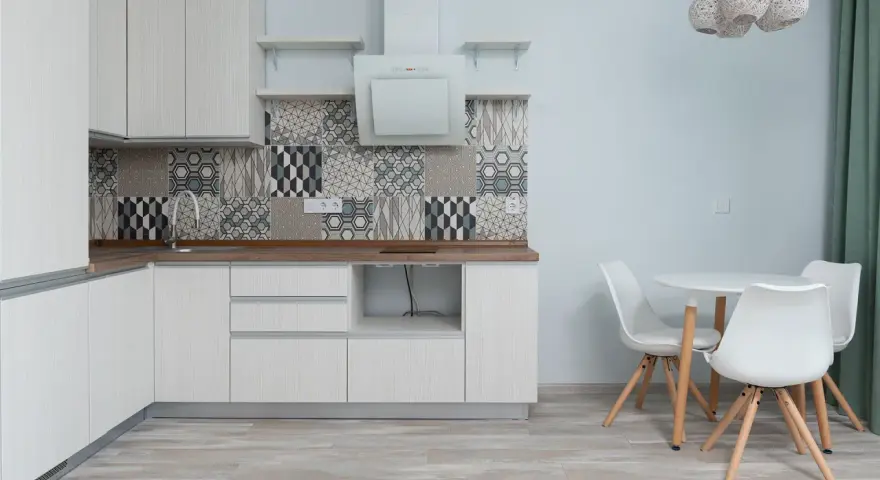 Cocina moderna con armarios blancos, salpicadero geométrico y suelo gris claro. Mesa redonda con sillas blancas.