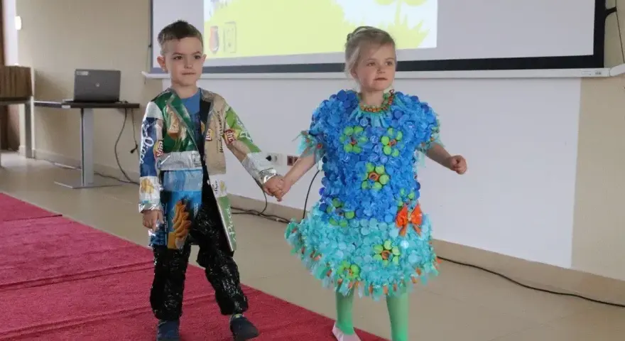 Chłopiec prezentuje strój z recyklingu, wykonany z opakowań po przekąskach. Jego koleżanka ma sukienkę z nakrętek.