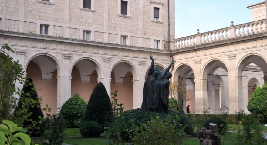 A bronze statue of monks with arms raised stands in a courtyard garden, perhaps commemorating St. Benedict's feast day.