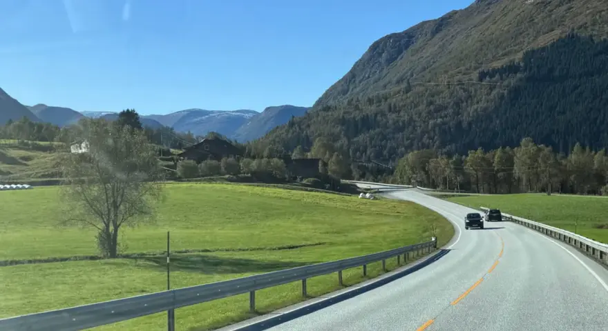 En bil kör på en slingrande väg genom ett grönt landskap med berg och hus. En perfekt start på en semester i Norge.