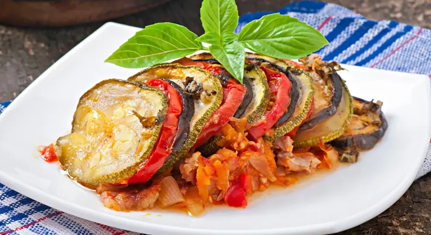 Ratatouille con rodajas de calabacín, berenjena y tomate, decorado con albahaca. Los ingredientes ratatouille se ven tiernos y jugosos.