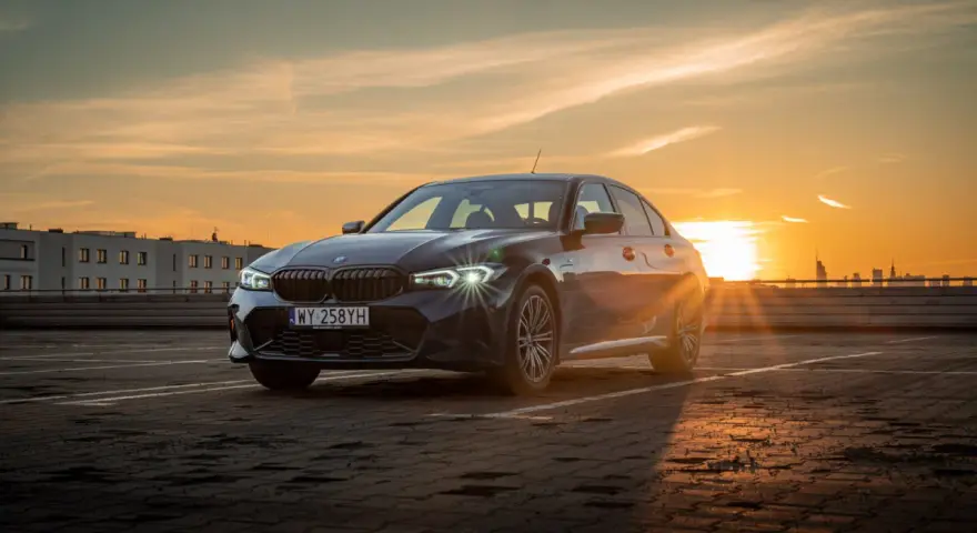 Ciemnoszara limuzyna BMW 330e na parkingu o zachodzie słońca.