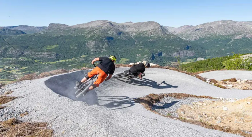Två cyklister kör utför en grusig bana i Hemsedal sommar. Berg och grönska i bakgrunden.