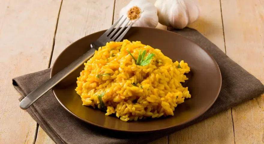Un invitante risotto zafferano in polvere, guarnito con prezzemolo fresco, su un piatto marrone, con aglio e forchetta sullo sfondo.