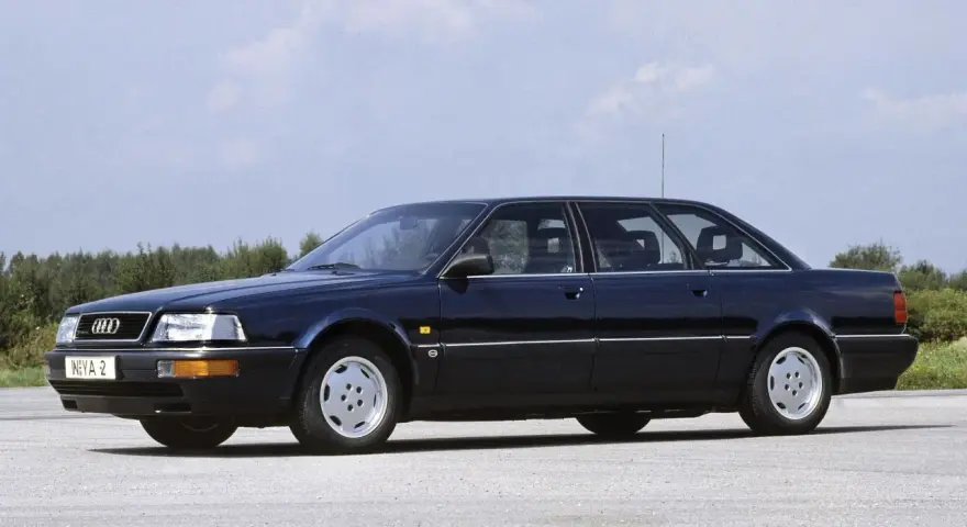 Ciemnoniebieski Audi V8 z lat 90. stoi na pustej drodze, z drzewami w tle.