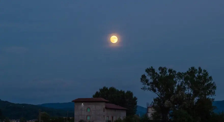 La luna di luglio illumina un paesaggio serale con colline, alberi e un edificio.