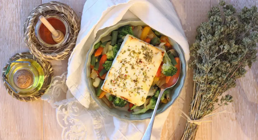 Feta al forno con verdure miste, condita con erbe aromatiche. Accanto, olio, spezie e un mazzetto di origano secco.